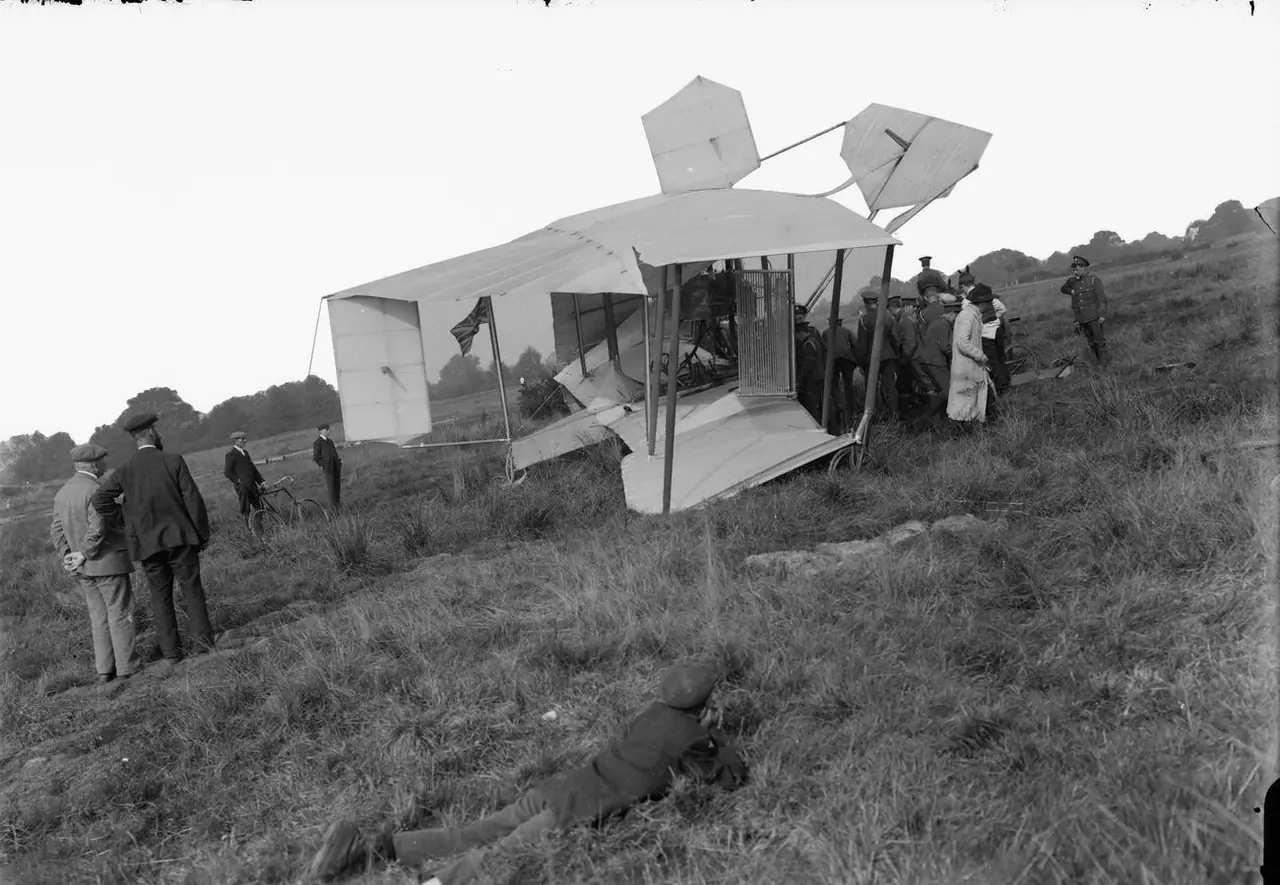 The wrecked British Army Aeroplane No 1 on open heathland after its flight, with a man in a white coat, onlookers and bicycles nearby