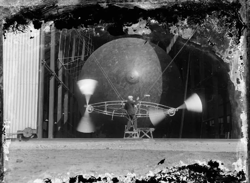 A man at the engine and spinning propellers of the Nulli Secundus airship in an open balloon shed at Farnborough, 1907